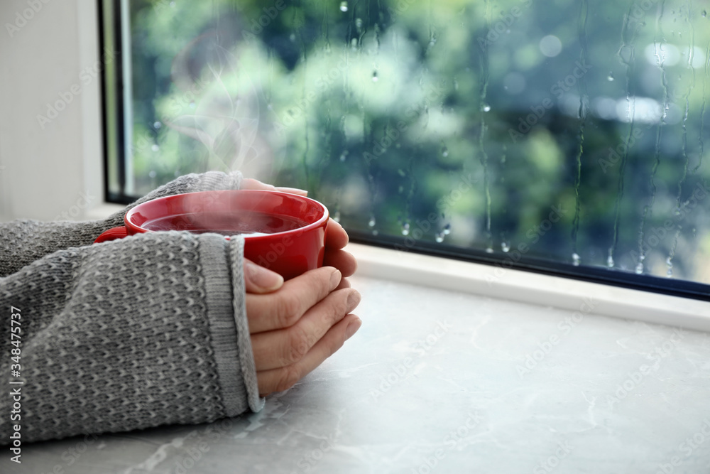 Woman with cup of hot drink near window on rainy day, closeup