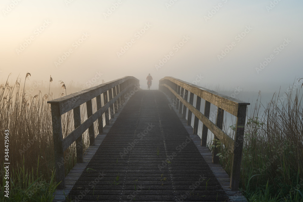 Cyclist on a wooden bridge over a river on a foggy morning in the dutch countryside.