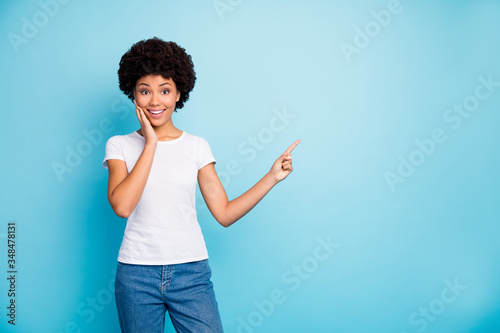 Photo of funny pretty dark skin curly lady sales manager indicating fingers empty space proposing low prices wear casual white t-shirt jeans isolated blue color background