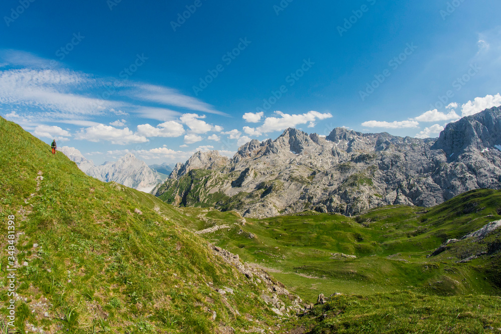 hiker in the middle of the mountain landscape, green meadows forming hills, gray limestone mountains background with blue sky
