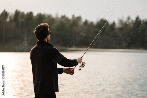 Fishing for pike, perch, carp. Fisherman with rod, spinning reel on river bank. Man catching fish, pulling rod while fishing on lake, pond. Wild nature. The concept of rural getaway. Back view.