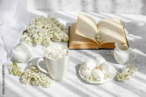 Still life vase with a bouquet of white lilac  a Cup of coffee with marshmallows  a plate of meringue  a sugar bowl and a milk jug   an old book. Postcard good morning.