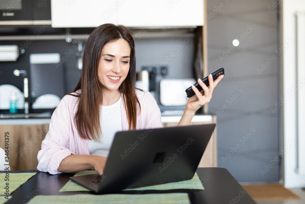 Young woman scroll phone while using laptop at table indoors Young woman scroll phone while using laptop at table indoors