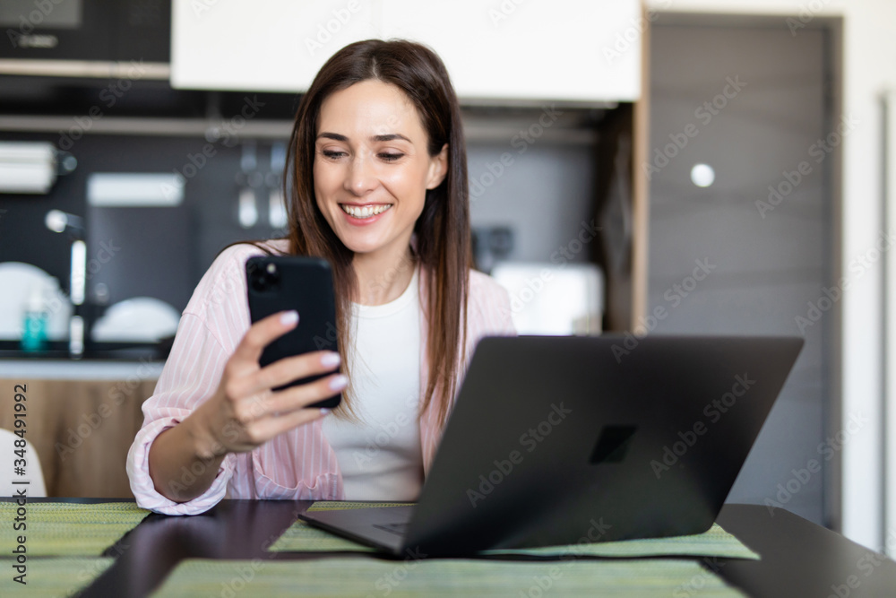 Young woman scroll phone while using laptop at table indoors Young woman scroll phone while using laptop at table indoors