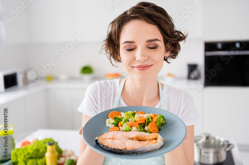 Close up photo of pretty housewife lady chef hold ready grilled salmon trout fillet steak with garnish cook dinner one person portion eyes closed wear apron t-shirt modern kitchen indoors photo
