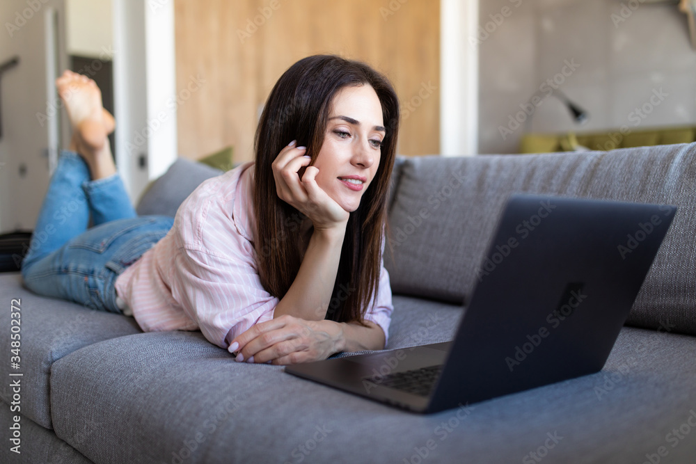 Beautiful woman smiling and relaxes with her laptop lying on her stomach on a sofa with her bare feet in the air Beautiful woman smiling and relaxes with her laptop lying on her stomach on a sofa with her bare feet in the air