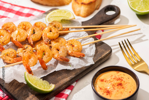 prawns on skewers with lime and sauce on parchment paper on wooden board and plaid napkin on white background