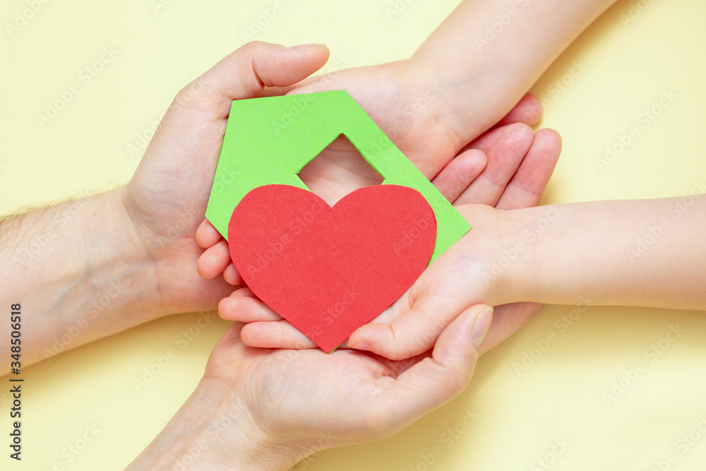 Close Up of dad and child hands holding green paper house with red heart on yellow background.