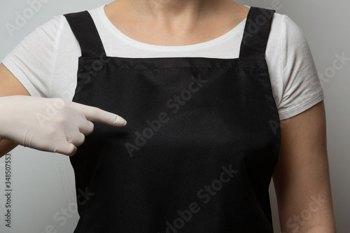 Closeup, fragment. Girl in black apron pointing finger to empty space on her chest. Protecting gloves. Safety concept. Copy space, place for text, mockup.