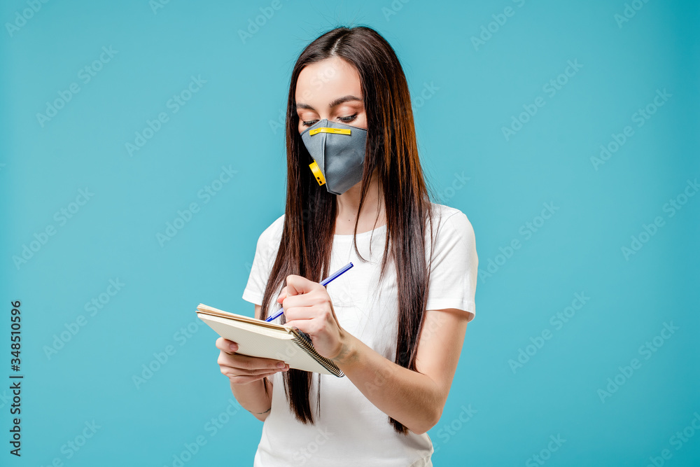 woman writing in notebook wearing respirator mask isolated on blue background