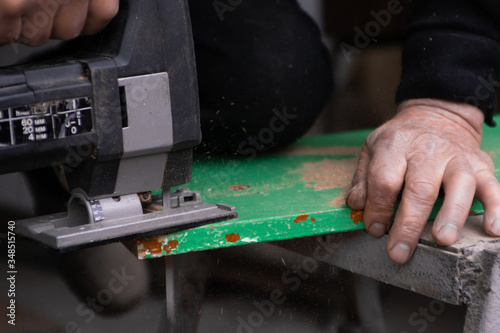 Mature man is working in garage with powered jigsaw and sawing a peace of green wooden plate