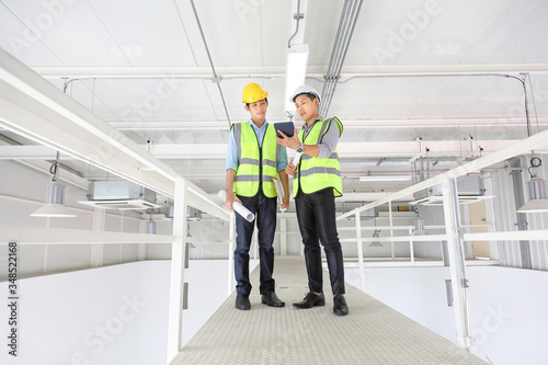 Two Asian contracting engineers wearing safety equipment while inspecting and discussing over the future expanding business plan of the factory inside the warehouse