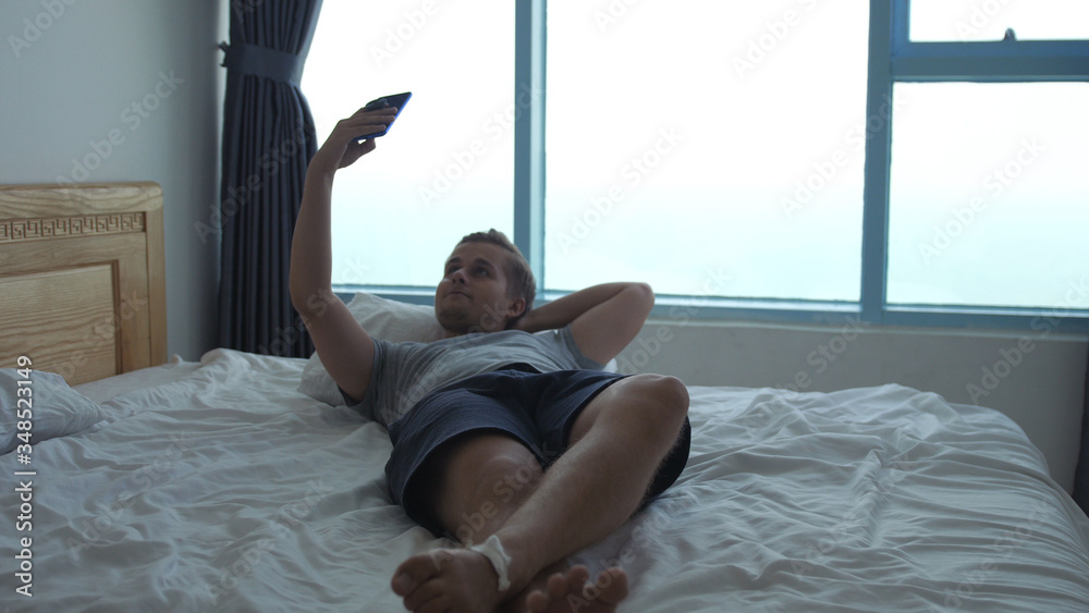 Young handsome man lies in bed by the window, uses the phone Young handsome man lies in bed by the window, uses the phone