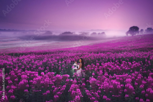 A beautiful girl in a pink dress standing with a lantern full of magic lights in a large pink field of willow herb. Romantic evening photo with sunset sky.
