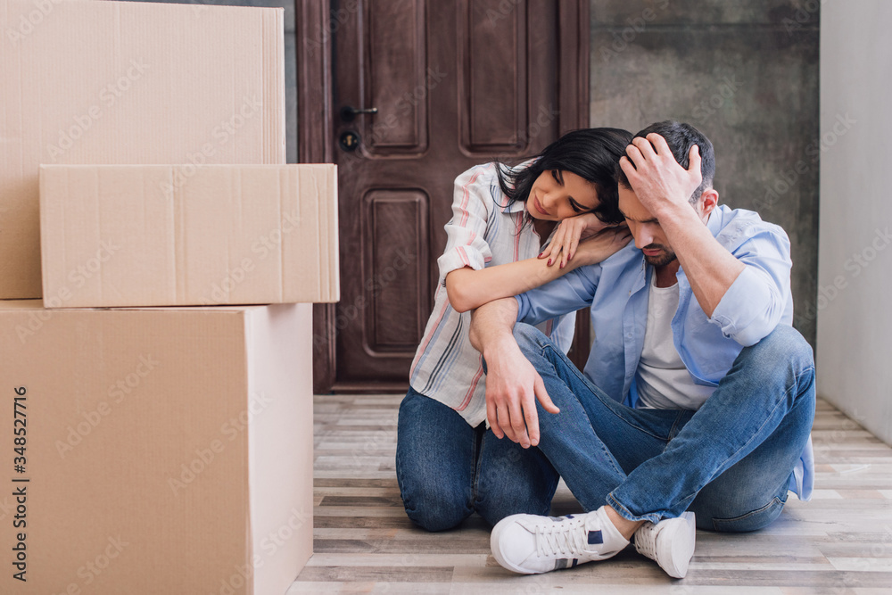 Upset woman leaning to husband with crossed legs near boxes on floor Upset woman leaning to husband with crossed legs near boxes on floor