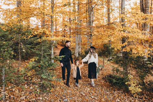 Stylish family in the autumn forest. A young guy and a girl are walking along a forest road among yellow leaves with their daughter.