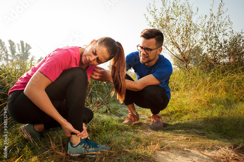 Worried young Caucasian man comforting his girlfriend who is experiencing serious ankle pain during early morning jogging in nature