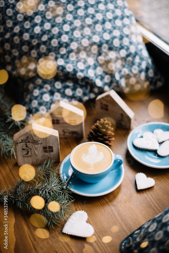 Delicious fresh cappuccino coffee in a blue cup with some heart shaped festive cookies, fireflies, spruce leaves and christmas decorations (little wooden houses) on the wooden table background © anastasianess