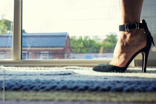 Woman feet wearing black highheels standing on carpet next to window in house over sky background.