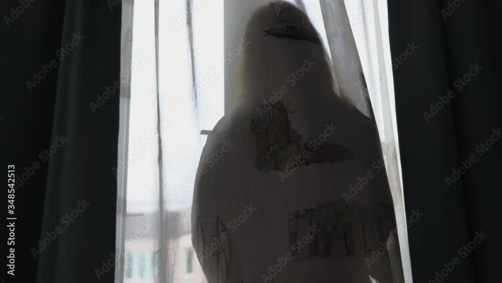 Silhouette of woman against window behind transparent curtain, woman in dressing gown looking back to room smiling while holding cellphone.