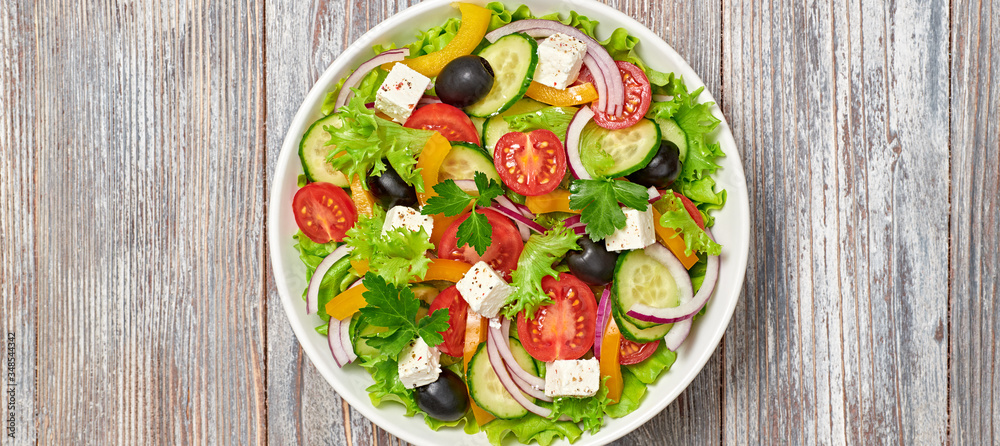 Greek salad with cucumeber, olives, feta cheese, cherry tomatoes, bell pepper and lettuce. Summer diet salad concept. Tasty greek salad in bowl on wood, top view, banner