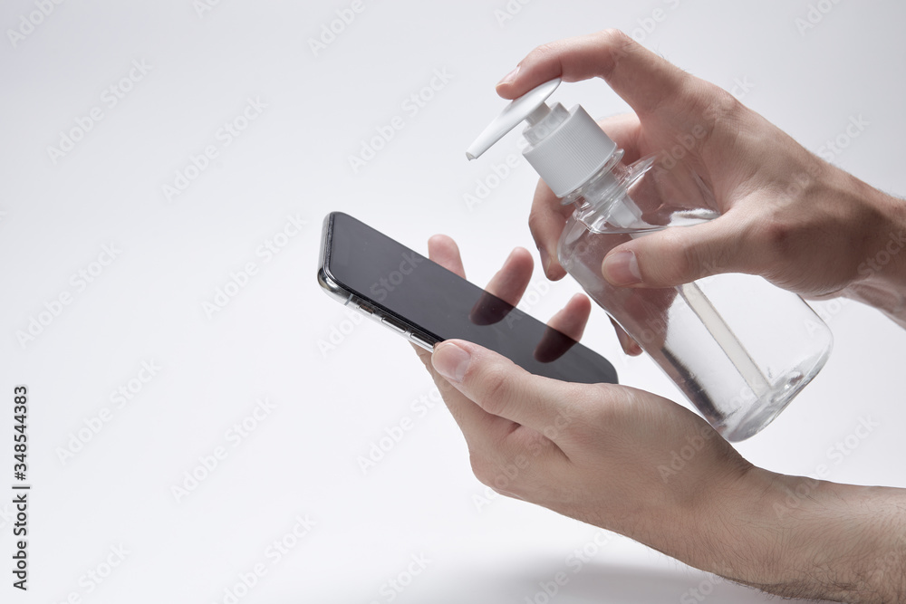 Fototapeta premium Disinfection of the phone. Hands hold smartphone and bottle or container for antiseptic cream or gel white background.
