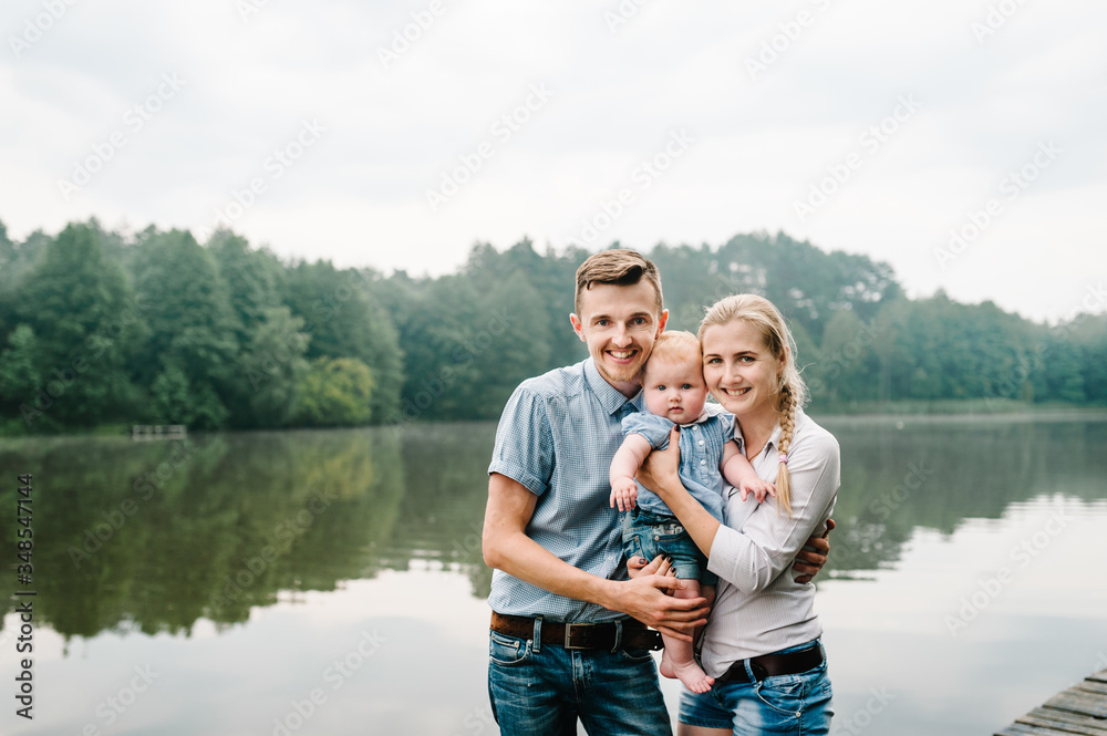 Happy young family near lake, pond. Family enjoying life together at meadow. People having fun in nature. Family bonds outside. Mother, father, child of three smiling while spending free time outdoors Happy young family near lake, pond. Family enjoying life together at meadow. People having fun in nature. Family bonds outside. Mother, father, child of three smiling while spending free time outdoors