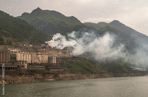 Xinling, China - May 6, 2010: Xiling gorge on Yangtze River. Smoke escaping from large cement plant along shoreline with green mountains in back under brown sky. bow bridge nearby. photo