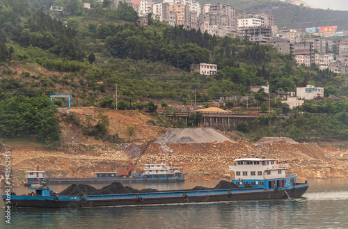 Xinling, China - May 6, 2010: Xiling gorge on Yangtze River. Coal barge sails on greenish water in front brown stone shoreline, where other barge is. On top, dense urban multi-story buildings and gree photo