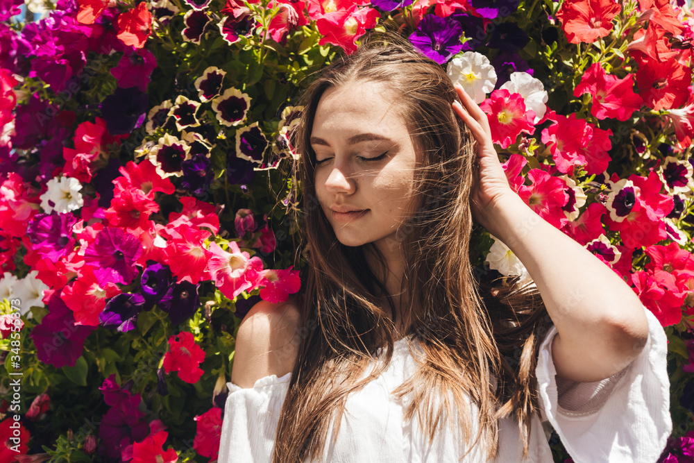 Naklejka premium covering her eyes a beautiful brunette straightens her hair against a background of blooming petunias