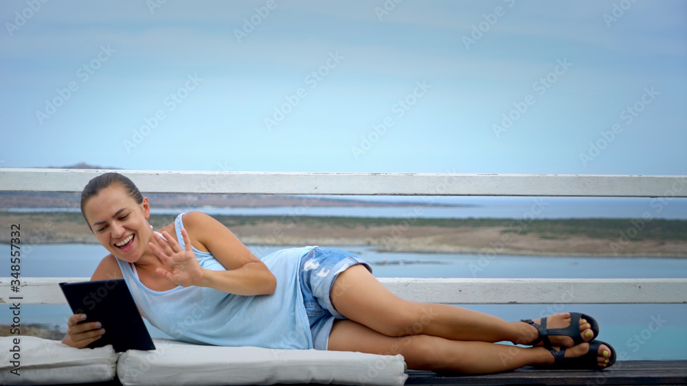 Beautiful woman talking on video chat while lying on the terrace by the sea
