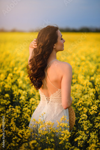 Summer mood. A young beautiful caucasian woman in a white dress is standing in a blooming yellow field back to camera. © MZaitsev