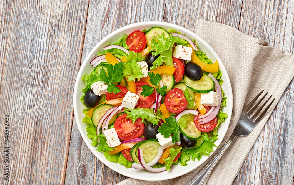 Greek salad with cucumeber, olives, feta cheese, cherry tomatoes, bell pepper and lettuce. Summer diet salad concept. Tasty greek salad in bowl on wood, top view