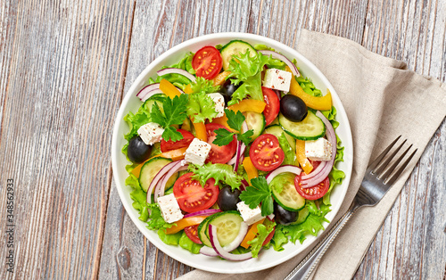 Greek salad with cucumeber, olives, feta cheese, cherry tomatoes, bell pepper and lettuce. Summer diet salad concept. Tasty greek salad in bowl on wood, top view