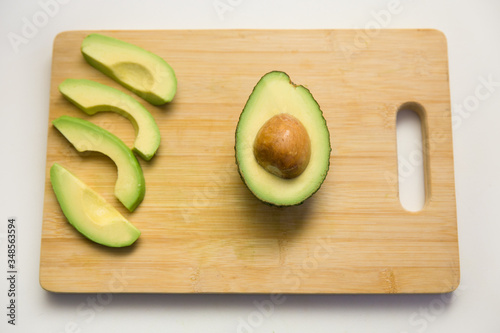 Parts of avocado fruit on wooden chopping board, half with seed and cut slices. Closeup shot, top view. Isolated objects on white background. Fresh food or healthy diet concept