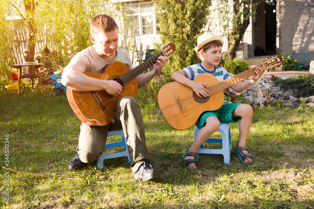 Fototapeta premium Happy family dad and child son having fun with musical instruments guitars together outdoors. Dad teaches son how to play guitar. quarantine. musical concept