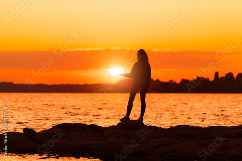 Girl standing on rock holding sun at hand. Nature and beauty concept. Orange sundown. Girl silhuette at sunset.