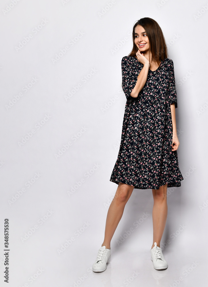 Brunette model in black dress with floral print, white sneakers. Smiling, touching her face, posing isolated on white. Full length Brunette model in black dress with floral print, white sneakers. Smiling, touching her face, posing isolated on white. Full length