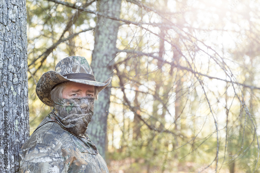 A middle aged man dressed for hunting leaning against a tree. He is wearing a camouflaged hat, shirt, and mask to blend in to the trees around him. The sun is beginning to set.