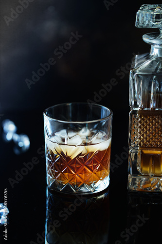 Glass of whiskey or bourbon with ice on black stone table. Glass of whiskey with ice and a square decanter. Glass of scotch whiskey