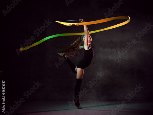 portrait of beautiful young brunette woman gymnast training calisthenics exercise with yellow ribbon on dark smoke studio background.