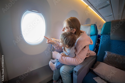 young beautiful mother sits in an airplane chair holds on her lap a little cute baby and points with finger into the porthole