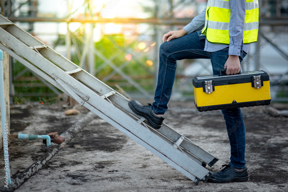Asian maintenance worker man with safety helmet and green vest carrying tool box climbing on aluminium ladder at construction site. Civil engineering, Architecture builder and building service concept Asian maintenance worker man with safety helmet and green vest carrying tool box climbing on aluminium ladder at construction site. Civil engineering, Architecture builder and building service concept