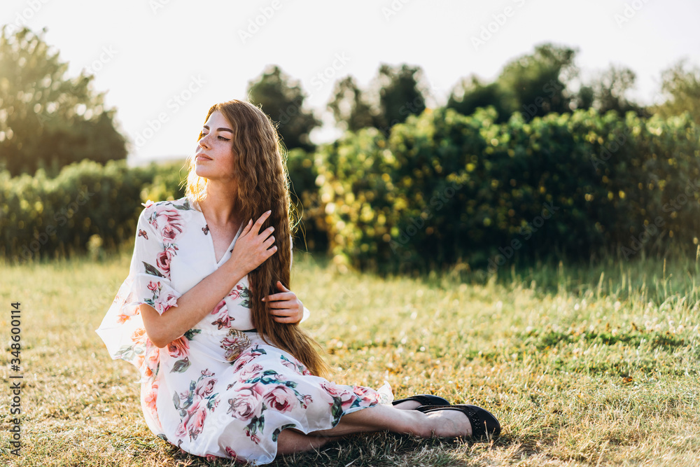 Full length portrait of beautiful woman with long curly hair on currant field background. Girl in a light dress sits on the grass in sunny day Full length portrait of beautiful woman with long curly hair on currant field background. Girl in a light dress sits on the grass in sunny day