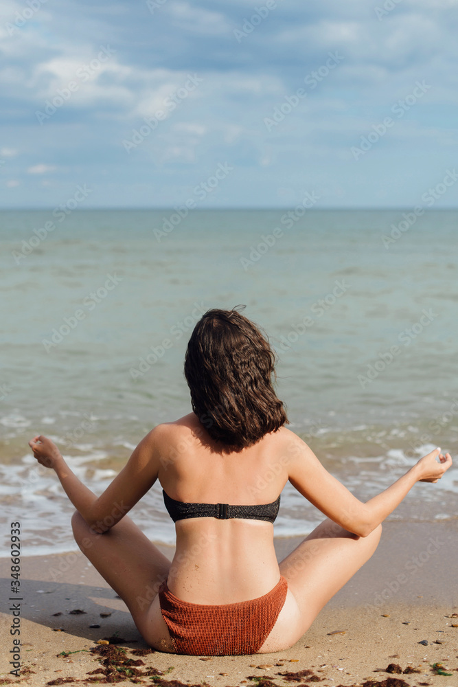 Young fit woman practicing yoga on the beach, sitting on sand and looking at sea waves. Girl meditating on summer vacation. Mental health and self care concept