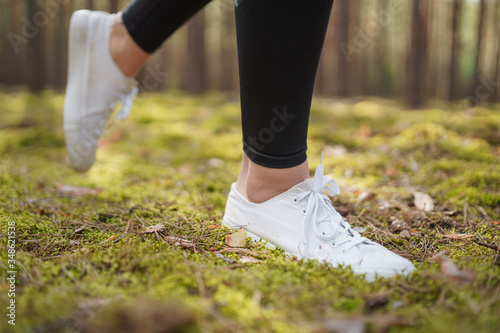 Runner feet running on road closeup on shoe. Jogging on a path in a pine forest. Woman running. White women's sneakers. Healthy lifestyle and weight loss concept.