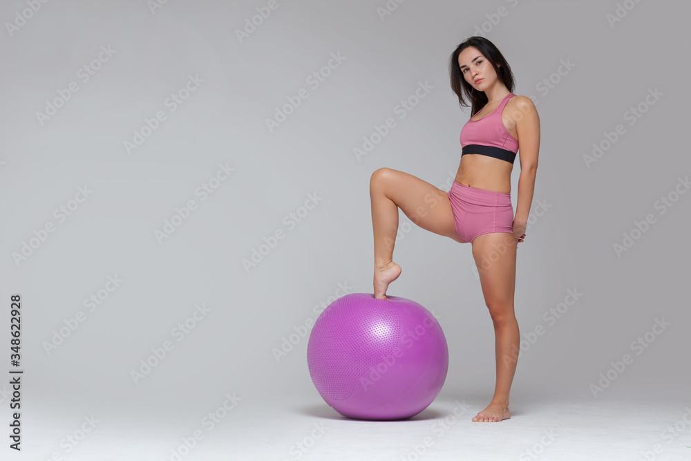 Young attractive girl in modern pink sportswear is posing putting her foot on fitness ball isolated on grey background. Full length Young attractive girl in modern pink sportswear is posing putting her foot on fitness ball isolated on grey background. Full length