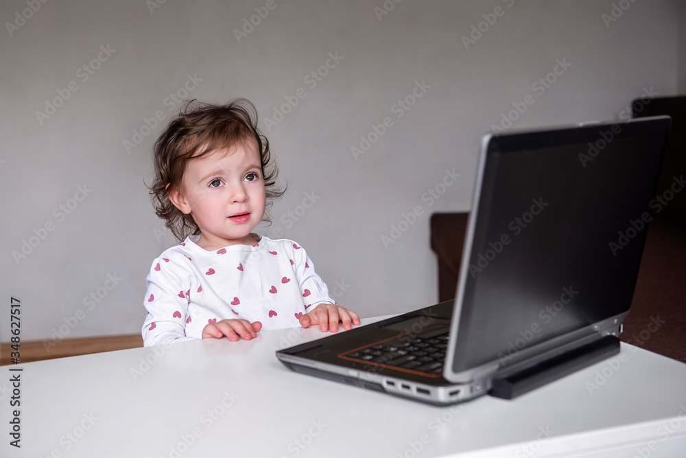 A charming little girl sits behind a laptop and watches and learns from a distance and participates in early development classes. Selective front and back focus, artistic noises. A charming little girl sits behind a laptop and watches and learns from a distance and participates in early development classes. Selective front and back focus, artistic noises.