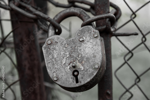 A close-up of an old locked iron lock with traces of paint on a thick chain wrapped between the gates of a metal fence on a gray gloomy background. A secret hidden from prying eyes. © Aleksandr Kondratov