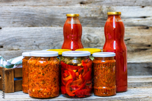 Glass jars with pickled red bell peppers and pickled cucumbers (pickles) isolated. Jars with variety of pickled vegetables, jars with zacusca and bottles with tomatoes sauce. Preserved food concept. photo